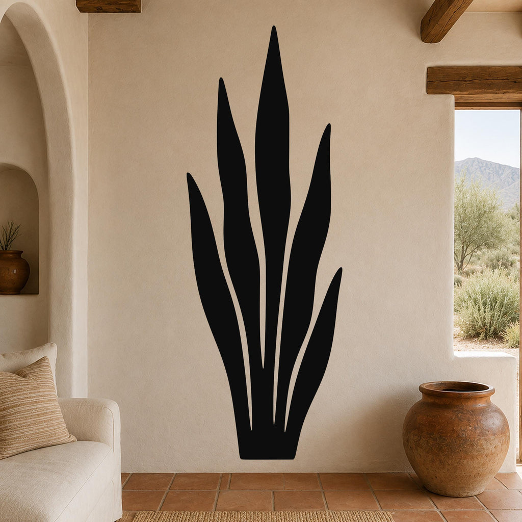 Black metal wall art in a tall, slender plant silhouette with internal cutout lines, mounted on a beige stucco wall in a living room with terracotta flooring and wooden beams.