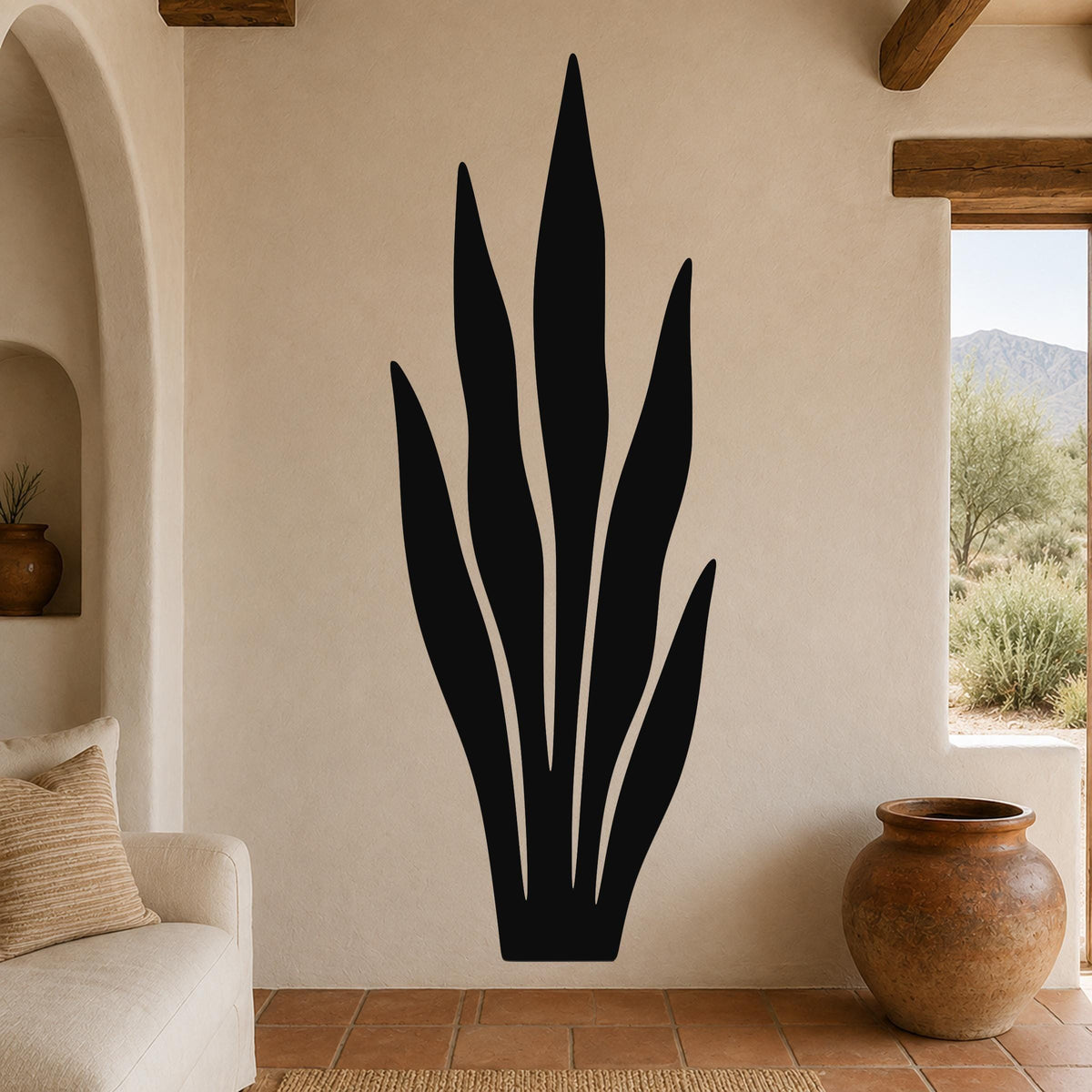 Black metal wall art in a tall, slender plant silhouette with internal cutout lines, mounted on a beige stucco wall in a living room with terracotta flooring and wooden beams.