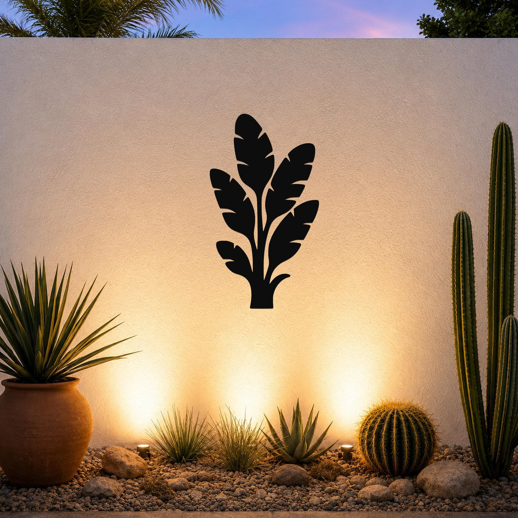 Black metal tropical leaves wall art silhouette mounted on a white outdoor wall above a gravel garden with cacti and warm uplighting.
