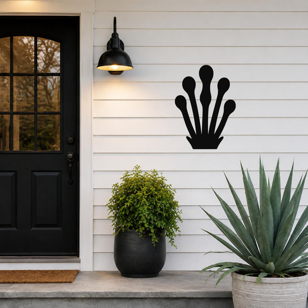 Black metal wall art in a silhouette of a kelp or seaweed frond with multiple rounded tips, mounted on a white horizontal siding wall beside a front door and potted plants.