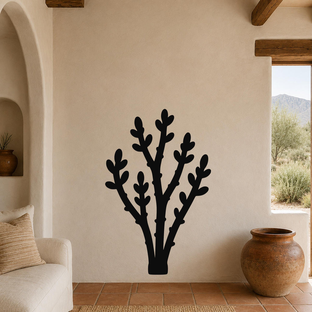 Black metal wall art in a desert branch silhouette, mounted on a beige stucco wall in a living room with terracotta flooring and wooden beams.