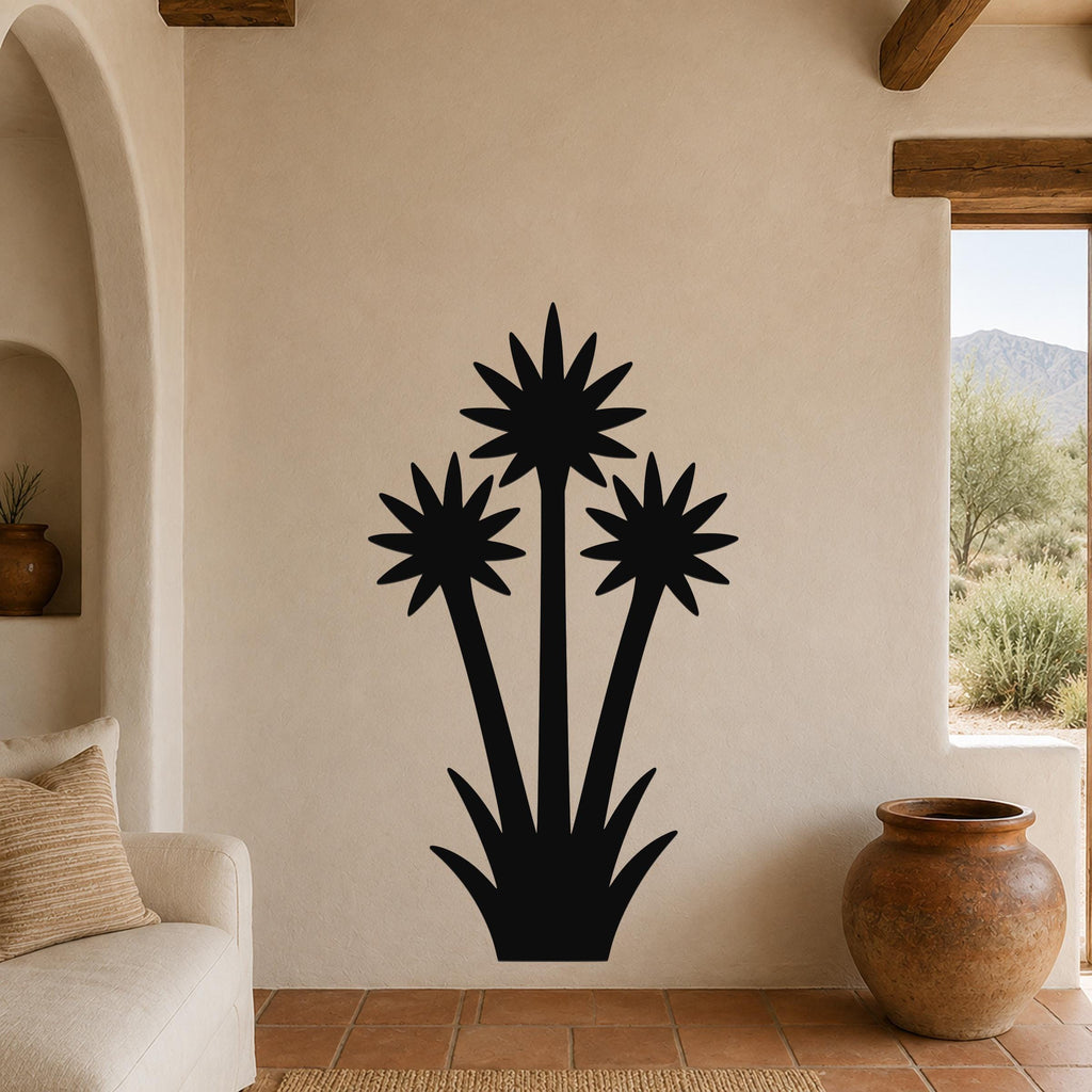 Black metal wall art silhouette of a blooming yucca plant with three star-shaped flower heads and spiky base, mounted on a beige stucco wall in a living room with terracotta tile floor and wooden beams.