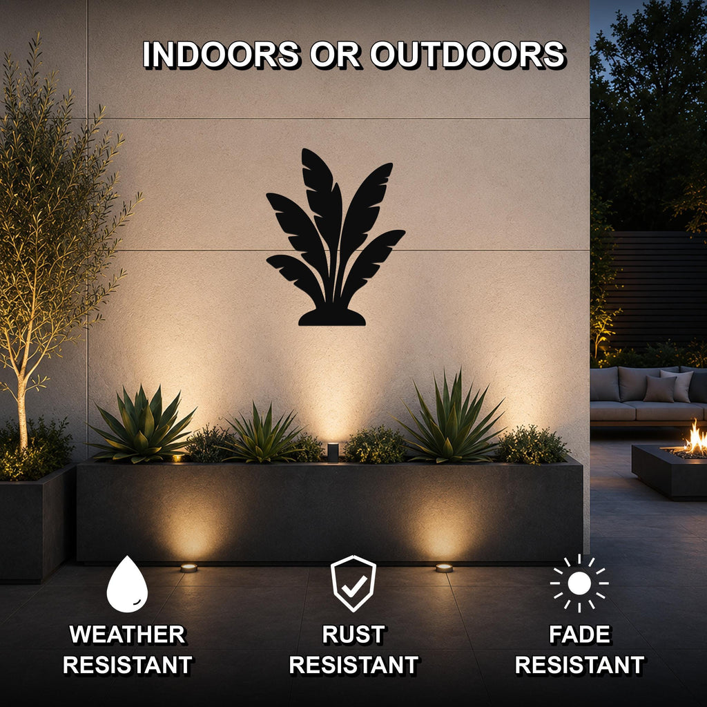 Black metal tropical leaves wall art silhouette mounted on an outdoor wall above a planter, illuminated by ground lights in a patio setting.