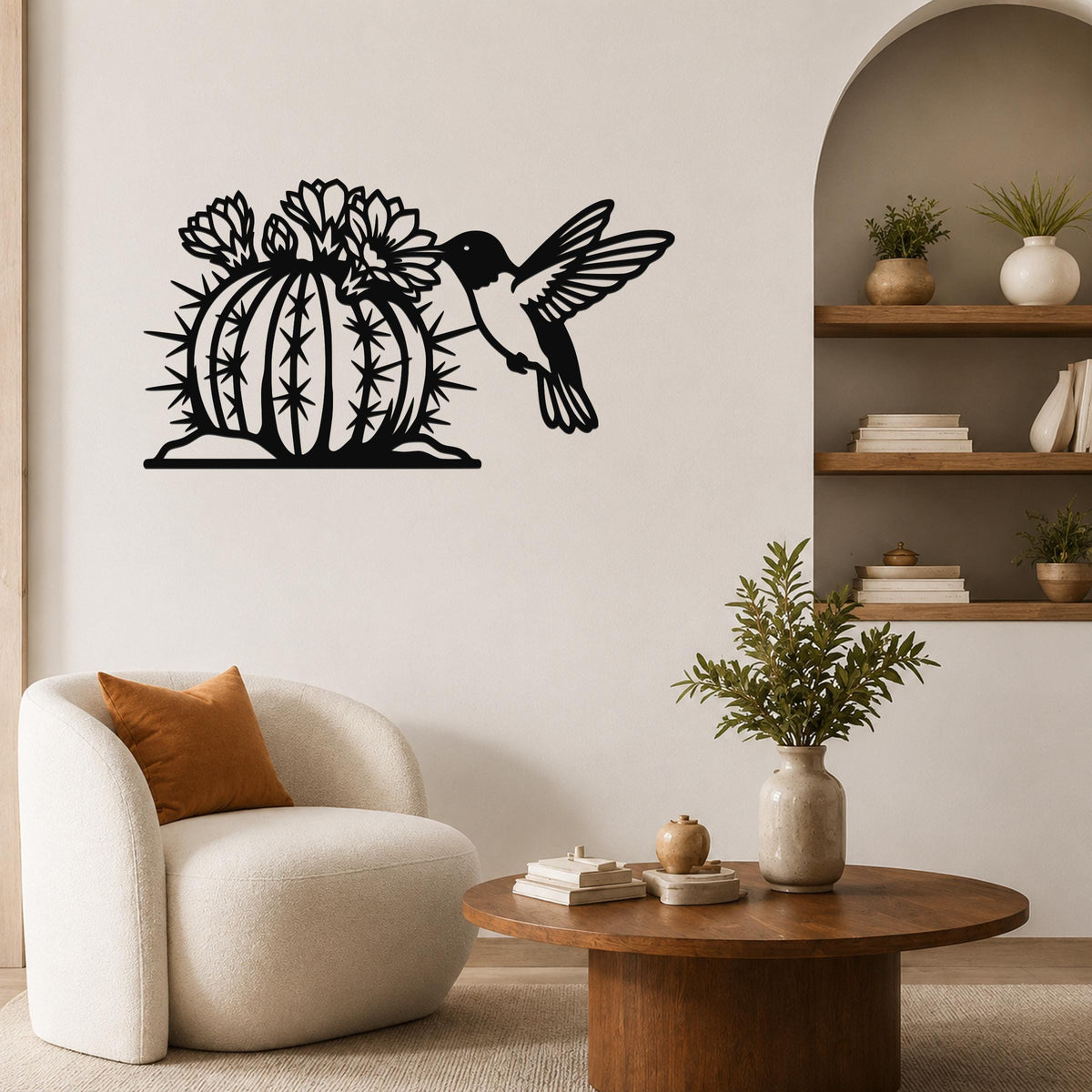 Black metal barrel cactus wall art with flowering blooms and a hummingbird cutout, mounted on a living room wall above a white armchair.