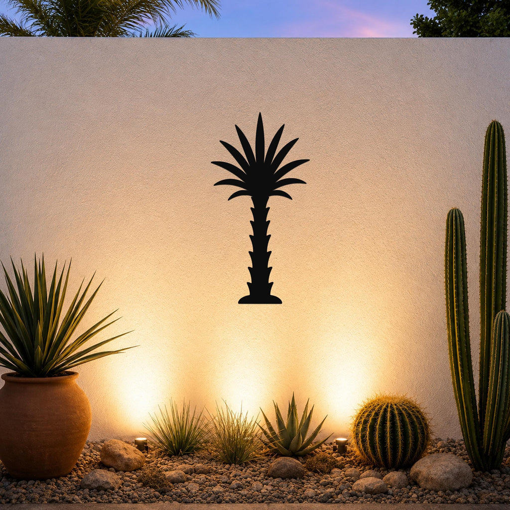 Black metal palm tree wall art silhouette mounted on a white outdoor wall above a desert garden with cacti and warm uplighting.