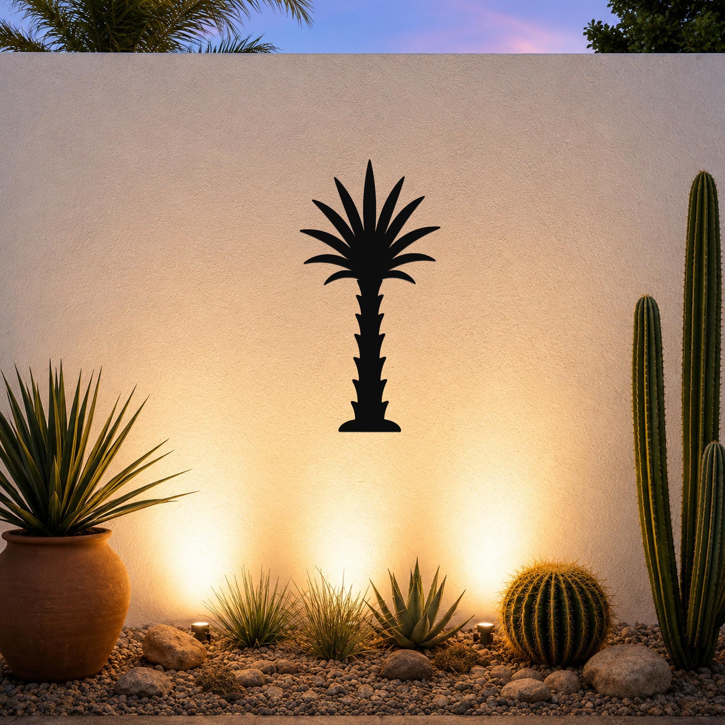 Black metal palm tree wall art silhouette mounted on a white outdoor wall above a desert garden with cacti and warm uplighting.