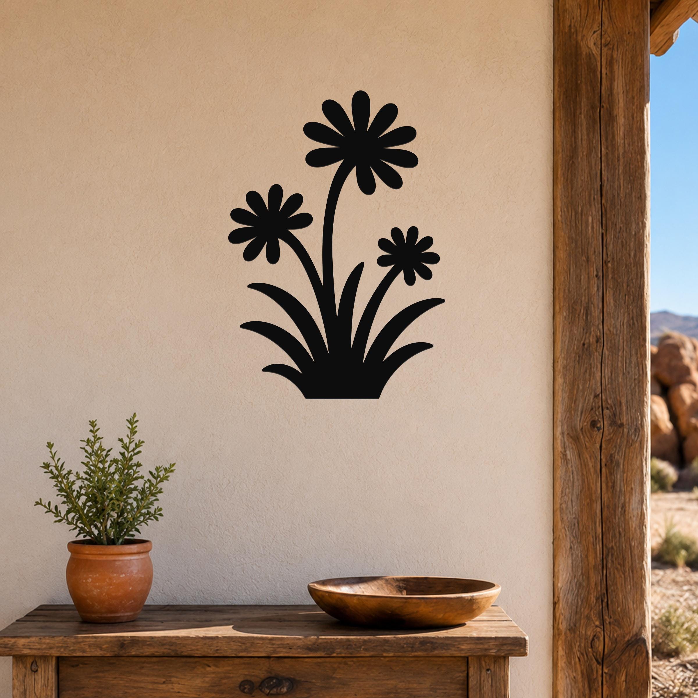 Black metal wall art featuring a silhouette of three daisies with long stems and leaves, mounted on a beige wall above a wooden table in a patio setting.
