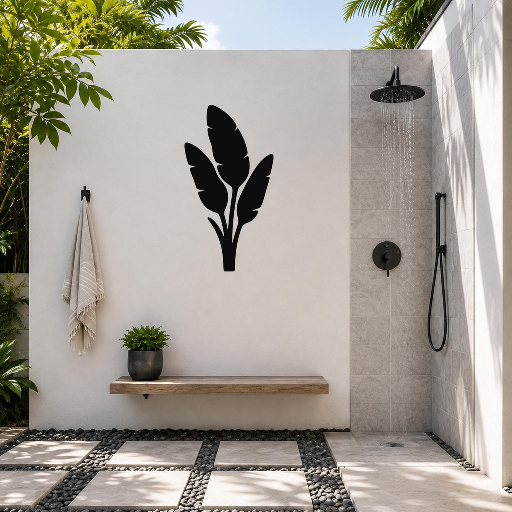 Black metal wall art with a tropical leaf silhouette, mounted on a white outdoor wall above a wooden bench in a patio shower area.