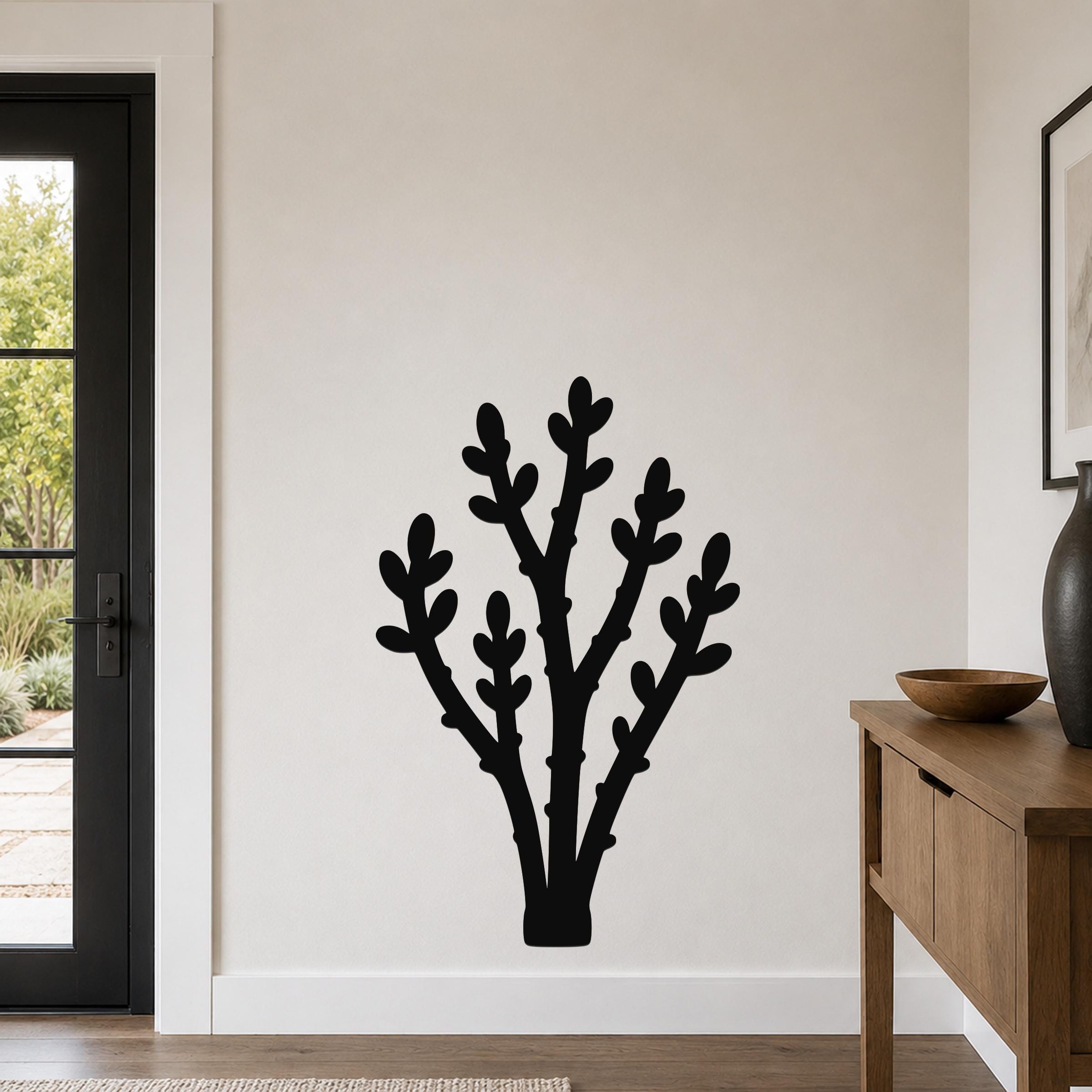 Black metal wall art in a silhouette of a desert branch with multiple stems and leaf cutouts, mounted on a light-colored wall in a living room near a door and console table.