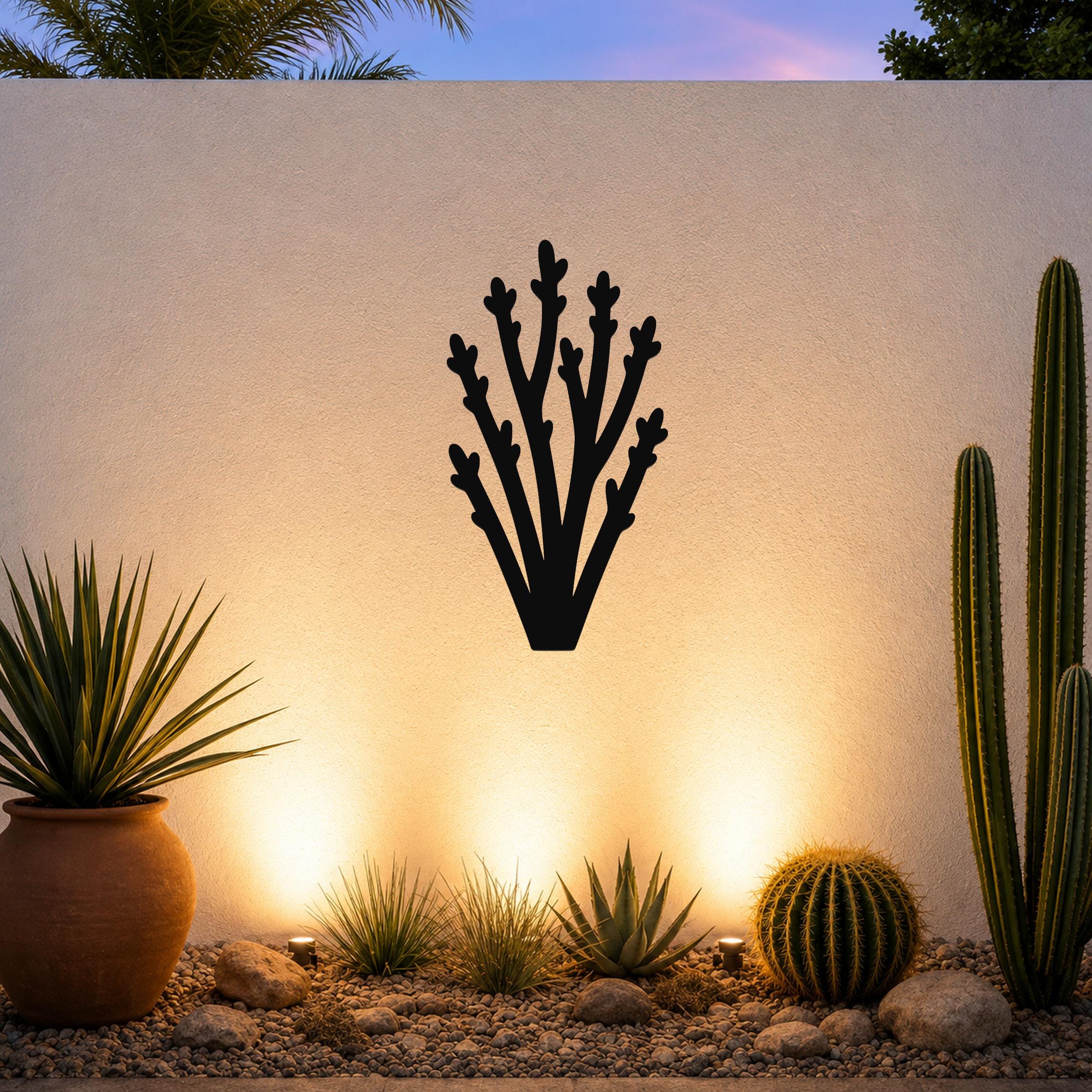 Black metal wall art in a desert branch silhouette, mounted on an outdoor wall above a gravel garden with cacti and warm uplighting.