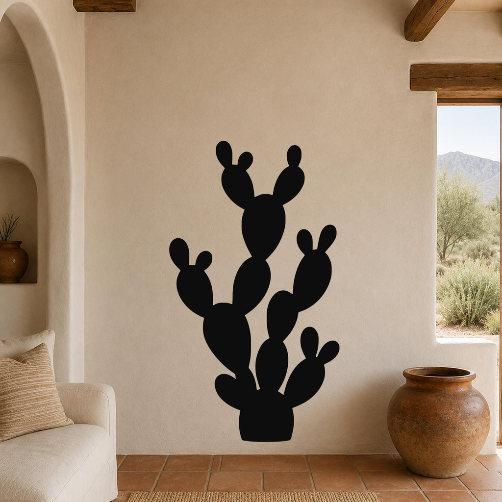 Black metal wall art in the silhouette of a prickly pear cactus, mounted on a beige stucco wall in a living room with terracotta flooring and wooden beams.