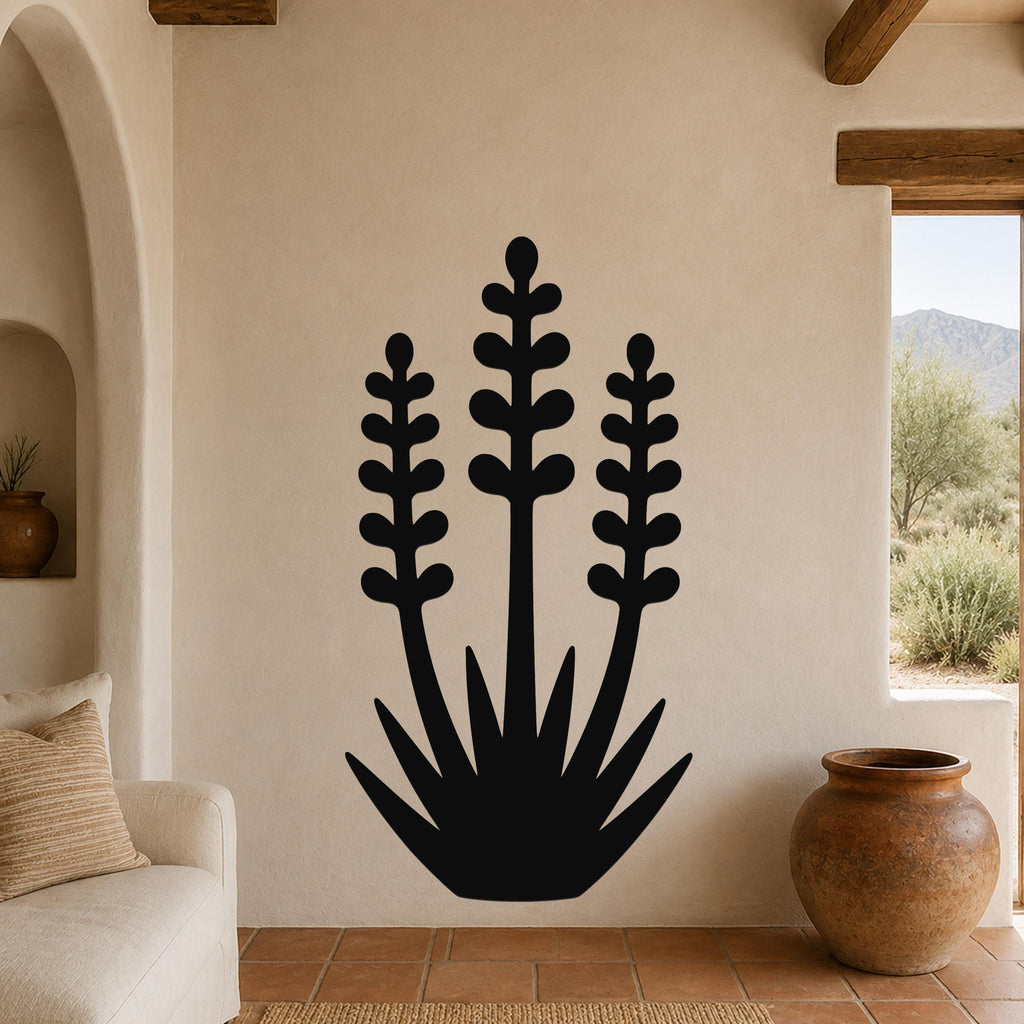 Black metal wall art in a yucca silhouette with three tall stems and spiky base, mounted on a beige wall in a living room with terracotta flooring and a view of mountains.