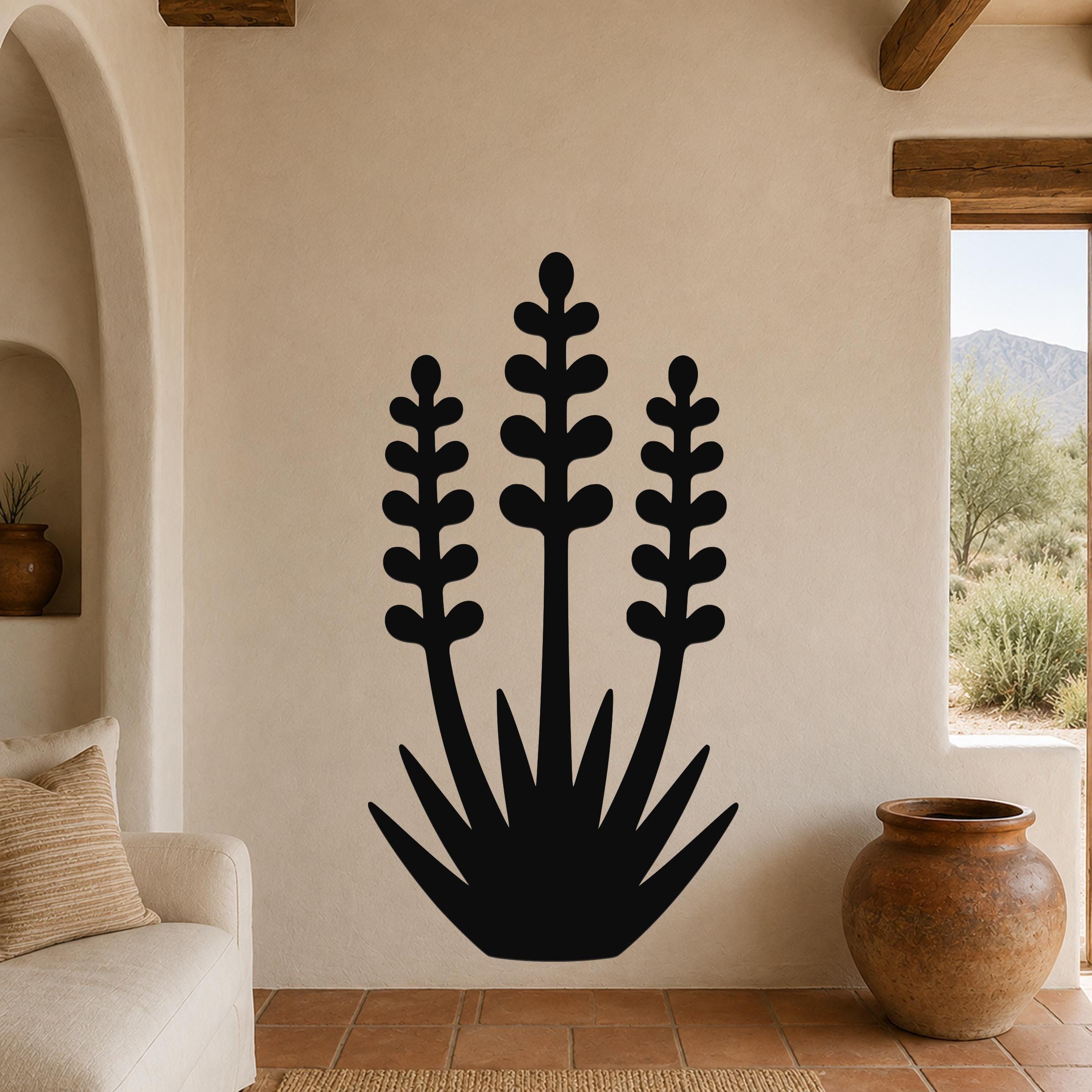 Black metal wall art in a yucca silhouette with three tall stems and spiky base, mounted on a beige wall in a living room with terracotta flooring and a view of mountains.